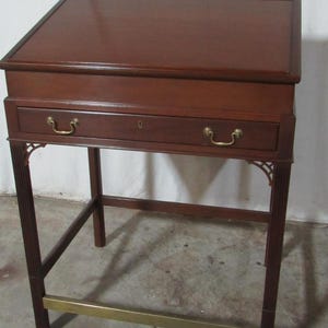 May include: A wooden writing desk with a slanted top and a drawer. The desk is a rich brown color with brass hardware and a brass bar connecting the legs. The desk has a classic, traditional design.