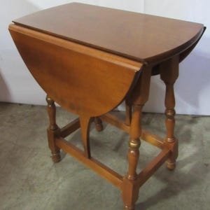 May include: A wooden drop-leaf table with a warm, medium-brown finish. The table features a rectangular top with rounded edges and a drop-leaf design. The legs are turned and connected by stretchers, creating a classic, traditional look.