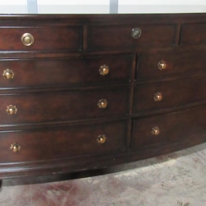 May include: A dark brown wooden dresser with nine drawers and ornate brass knobs. The dresser has a curved front and decorative carved details on the sides. A classic piece of bedroom furniture.