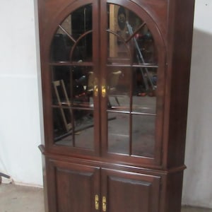 May include: A dark wood corner cabinet with glass doors and a lower cabinet. The upper doors have an arched top with a grid pattern, and the lower doors are solid. Gold-colored hardware accents the doors.