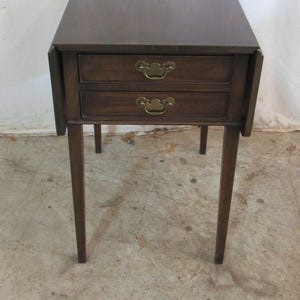 May include: A dark brown wooden end table with two drawers and brass handles. The table has four slender legs and a square top.