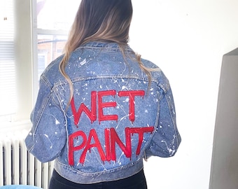 Könnte beinhalten: Hellblaue Jeansjacke mit weißen Farbspritzern und dem roten Schriftzug "WET PAINT" auf dem Rücken. Die Jacke wird mit einer schwarzen Hose getragen.