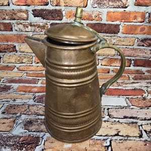 May include: A vintage brass coffee pot with a lid and handle. The pot has horizontal ridges and a spout for pouring. The handle is curved, and the lid has a small knob. The pot is set against a brick wall.