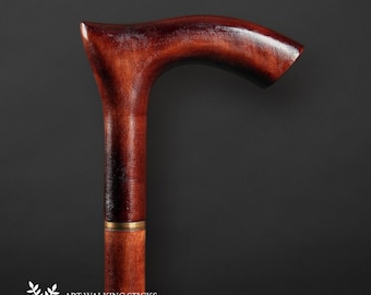 May include: A close-up of the top of a wooden walking stick with a curved handle. The handle is a rich reddish-brown color and has a brass band around it. The stick is made by Art Walking Sticks, a company that makes handmade products.