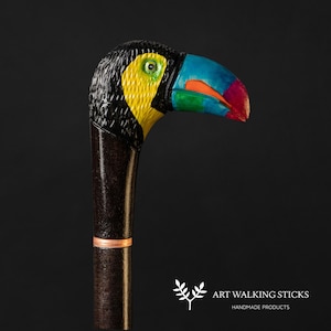 Può includere: Bastone da passeggio decorativo con testa di tucano. La testa del tucano è dipinta di nero, giallo, blu, verde e rosso. L'impugnatura è marrone scuro con una fascia color rame. Il testo "ART WALKING STICKS HANDMADE PRODUCTS" è visibile.