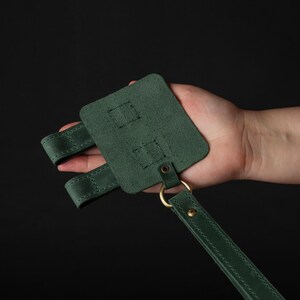 May include: A forest green leather accessory with a rectangular base, two straps, and a long strap. The accessory features brass hardware, including a ring and rivets. The item is held against a black background.