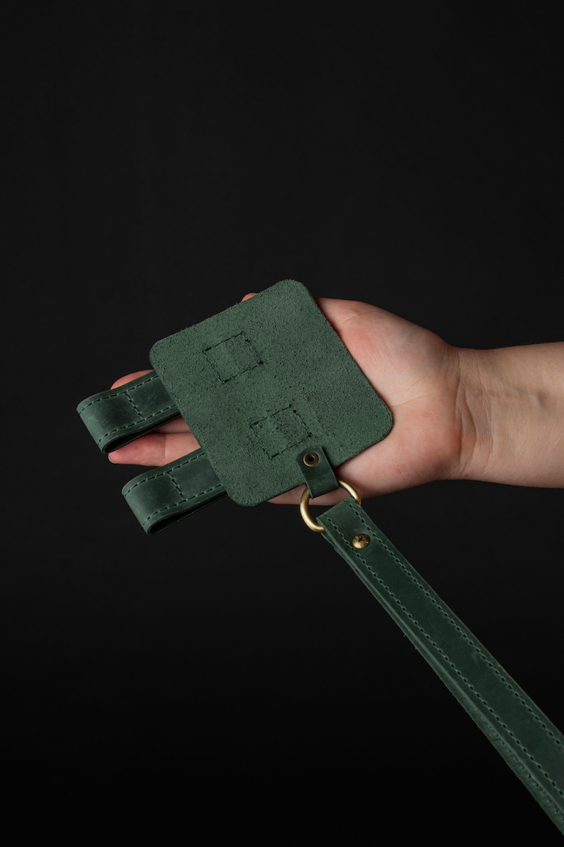 May include: A forest green leather accessory with a rectangular base, two straps, and a long strap. The accessory features brass hardware, including a ring and rivets. The item is held against a black background.