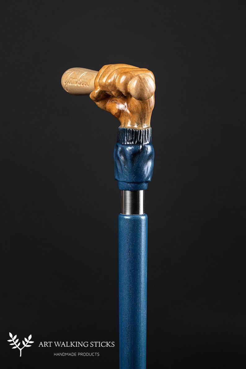 May include: A wooden walking stick with a hand holding a baseball bat. The hand and bat are carved from wood and painted in a natural wood tone. The stick is painted a dark blue color.