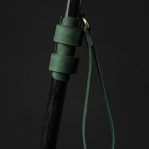 May include: A close-up of a walking cane with a dark wooden shaft and a decorative handle with gold and white accents. A green leather strap and handle are attached to the cane with a gold ring.