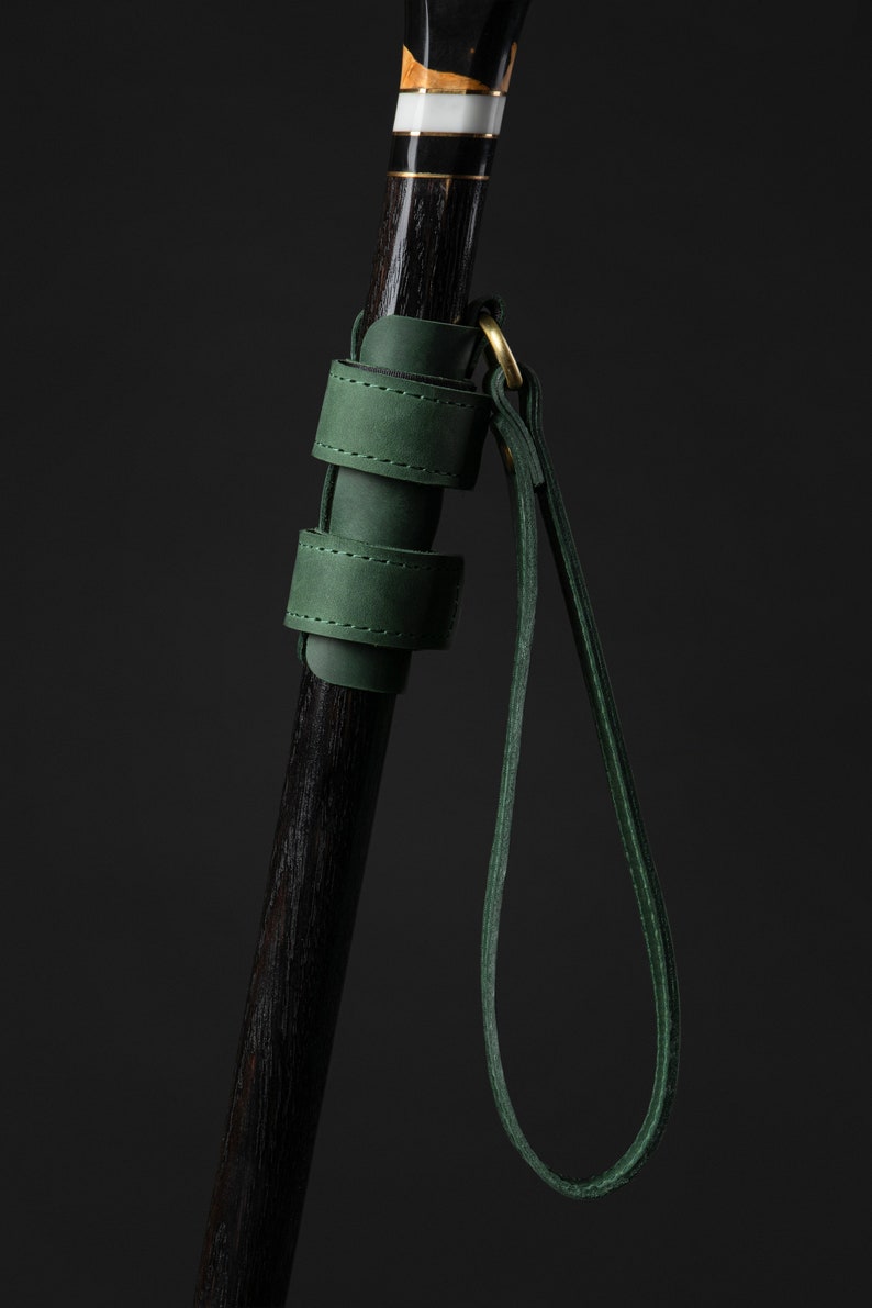May include: A close-up of a walking cane with a dark wooden shaft and a decorative handle with gold and white accents. A green leather strap and handle are attached to the cane with a gold ring.