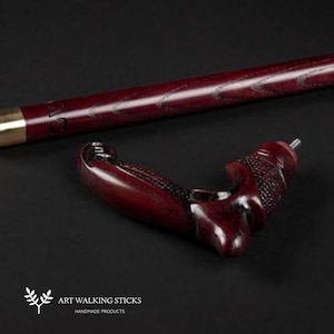 May include: A close-up of a dark red wooden walking stick with a carved handle. The handle is shaped like a hand with a detailed design. The stick is made by Art Walking Sticks, a company that makes handmade products.