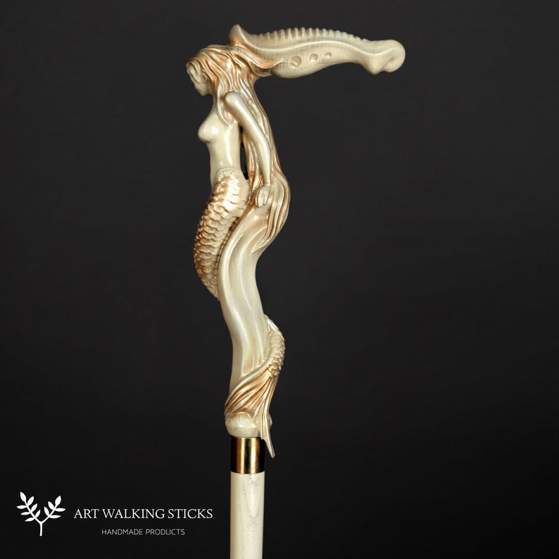 Puede incluir: Un bast&oacute;n de madera blanco y dorado con un dise&ntilde;o de sirena. La sirena est&aacute; tallada en detalle y tiene una cola fluida. El bast&oacute;n est&aacute; hecho de madera y tiene una punta de metal dorada.