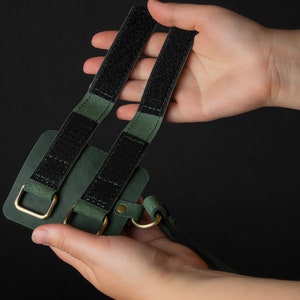 May include: A dark green leather phone case with black velcro straps and gold-colored metal hardware. The case has a loop for a strap and is held against a black background.