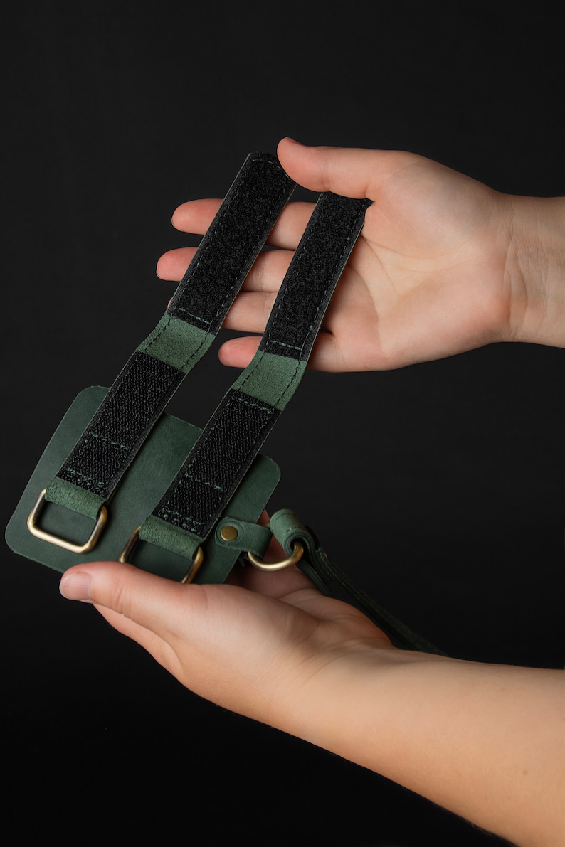 May include: A dark green leather phone case with black velcro straps and gold-colored metal hardware. The case has a loop for a strap and is held against a black background.