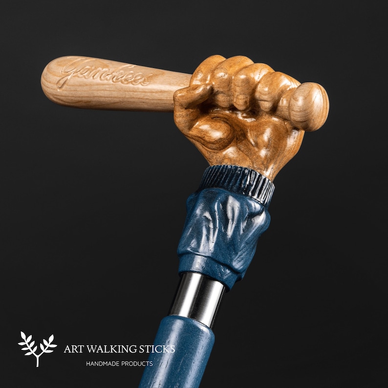 May include: A wooden walking stick with a carved baseball bat and hand. The hand is holding the bat and is wearing a blue sleeve. The bat is inscribed with the word "Yankees".