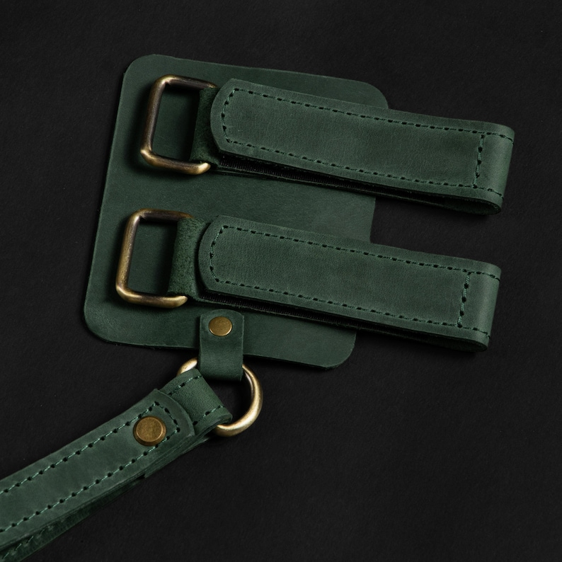 May include: Close-up of a dark green leather apron detail with brass-colored hardware. The apron features two adjustable straps with buckles and a connecting strap with a metal ring and rivet.
