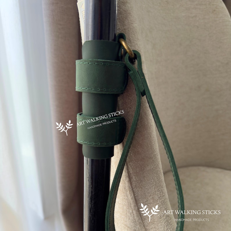 May include: A dark green leather walking stick holder with a matching strap and brass ring. The holder is attached to a dark wooden walking stick. The text "ART WALKING STICKS HANDMADE PRODUCTS" is visible.