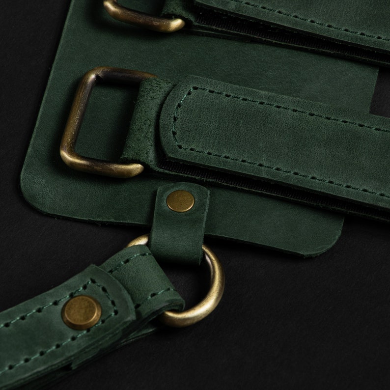 May include: Close-up of a dark green leather apron with brass-colored hardware. The apron features adjustable straps, metal buckles, and rivets. The leather has a textured appearance, and the stitching is visible.