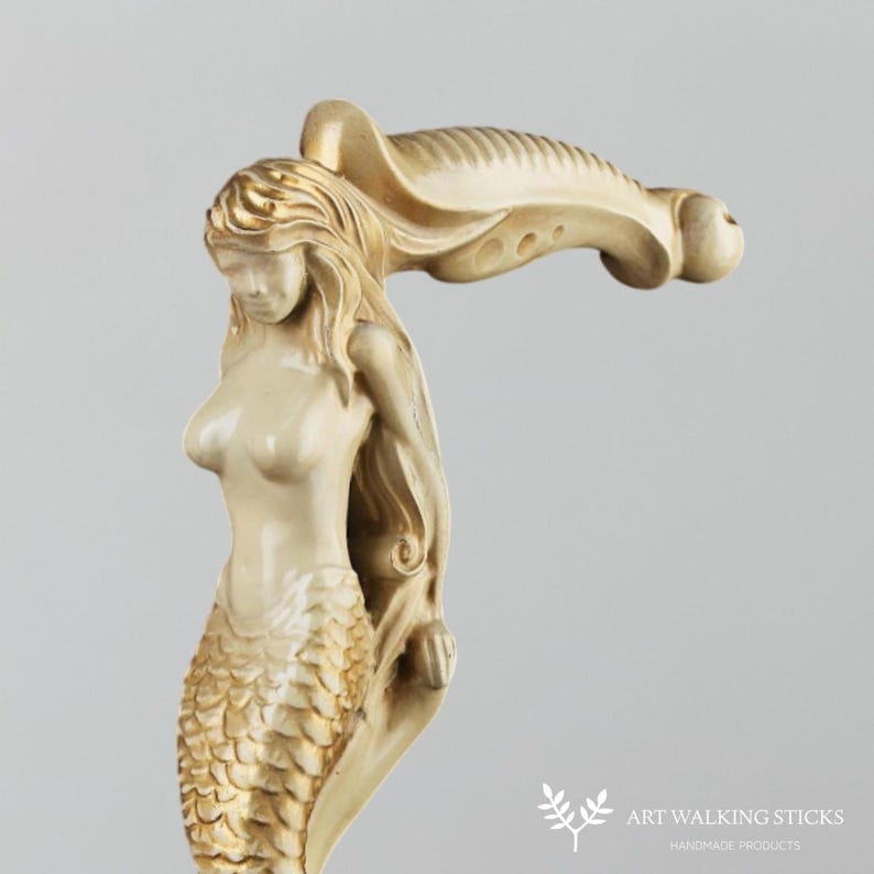 Puede incluir: Una escultura de sirena de color dorado y marfil con una cola fluida y un mango en forma de concha. La escultura es un mango de bast&oacute;n.