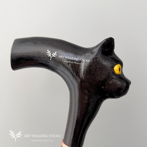 May include: Black cat-shaped walking stick with yellow eyes. The stick is made of wood and has a smooth, polished finish. The handle is shaped like a cat's head and has a small, white logo on the side that reads "Art Walking Sticks Handmade Products".