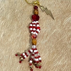 Pode incluir: Um chaveiro artesanal com um anel dourado e um pendão decorativo. O pendão apresenta contas vermelhas, brancas e transparentes, com detalhes dourados e um berloque de raio de latão. O chaveiro é exibido em uma superfície marrom clara.