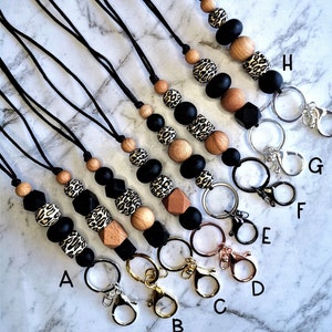 May include: A selection of beaded lanyards with black cords and metal clasps. The lanyards incorporate black, wood, and leopard print beads. The clasps are in silver, gold, and rose gold colours.