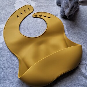 Puede incluir: Babero de silicona para beb&eacute; de color mostaza con cierre de cuello ajustable. El babero tiene un dise&ntilde;o curvo con un bolsillo para recoger los derrames. La palabra "MUSTARD" est&aacute; impresa en la parte inferior. Un conejito de peluche gris est&aacute; en el fondo.
