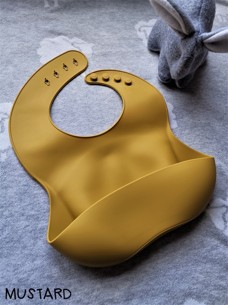 Puede incluir: Babero de silicona para beb&eacute; de color mostaza con cierre de cuello ajustable. El babero tiene un dise&ntilde;o curvo con un bolsillo para recoger los derrames. La palabra "MUSTARD" est&aacute; impresa en la parte inferior. Un conejito de peluche gris est&aacute; en el fondo.