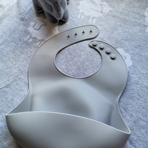 Puede incluir: Un babero de silicona gris claro con cierre de cuello ajustable. El babero tiene un bolsillo curvo en la parte inferior para recoger la comida. Un conejito de peluche gris est&aacute; en el fondo. El texto "LIGHT GREY" est&aacute; en la esquina superior derecha.