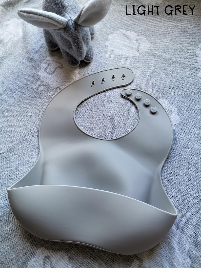 Puede incluir: Un babero de silicona gris claro con cierre de cuello ajustable. El babero tiene un bolsillo curvo en la parte inferior para recoger la comida. Un conejito de peluche gris est&aacute; en el fondo. El texto "LIGHT GREY" est&aacute; en la esquina superior derecha.