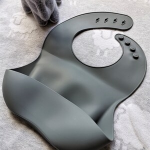 Puede incluir: Un babero de silicona gris pizarra con un dise&ntilde;o curvo y un bolsillo para recoger la comida. El babero tiene broches ajustables y un cuello redondeado. La palabra "SLATE" est&aacute; impresa en negro en la esquina inferior derecha. Un peluche gris est&aacute; en el fondo.