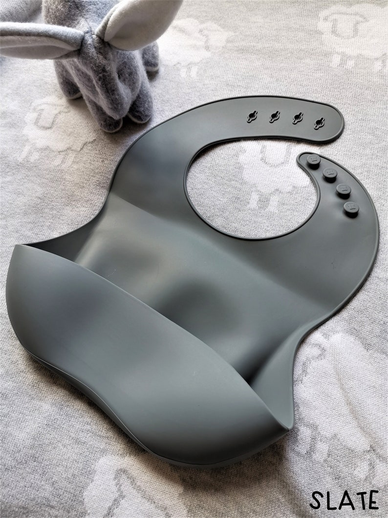Puede incluir: Un babero de silicona gris pizarra con un dise&ntilde;o curvo y un bolsillo para recoger la comida. El babero tiene broches ajustables y un cuello redondeado. La palabra "SLATE" est&aacute; impresa en negro en la esquina inferior derecha. Un peluche gris est&aacute; en el fondo.
