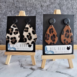 May include: Two pairs of leopard print dangle earrings with black teardrop-shaped leather and black round posts. The earrings are displayed on small wooden easels with a black card that reads "BIG BEAR AND LITTLE BIRDIE".
