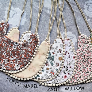 Puede incluir: Seis baberos para bebé con diferentes estampados y pompones blancos alrededor de los bordes. Los baberos están etiquetados como "Arlo", "Poppy", "Marely", "Sage", "Willow" y "Hazel".