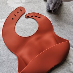 Puede incluir: Un babero de silicona color &oacute;xido para beb&eacute; con cuello ajustable y bolsillo curvo. El babero tiene una superficie lisa y est&aacute; dise&ntilde;ado para una f&aacute;cil limpieza. La palabra "AUTUMN" est&aacute; impresa en la parte inferior.