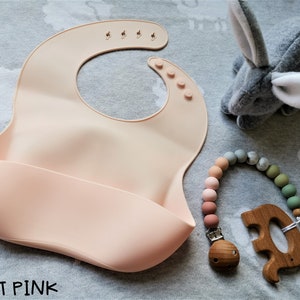 Puede incluir: Un babero de silicona rosa claro con un bolsillo para atrapar alimentos, un mordedor de madera y un clip para chupete con cuentas. Un conejito de peluche gris tambi&eacute;n est&aacute; en la imagen. El texto "LIGHT PINK" est&aacute; en la parte inferior de la imagen.