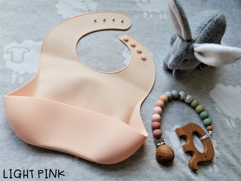 Puede incluir: Un babero de silicona rosa claro con un bolsillo para atrapar alimentos, un mordedor de madera y un clip para chupete con cuentas. Un conejito de peluche gris tambi&eacute;n est&aacute; en la imagen. El texto "LIGHT PINK" est&aacute; en la parte inferior de la imagen.