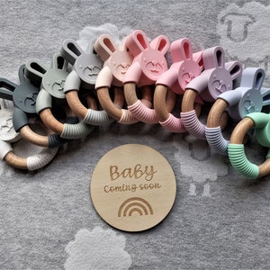 Puede incluir: Un conjunto de nueve anillos de dentición de madera con cabezas de conejito de silicona en varios colores pastel. Los anillos están dispuestos en un semicírculo con un letrero de madera que dice "Baby Coming Soon" y un gráfico de arcoíris.