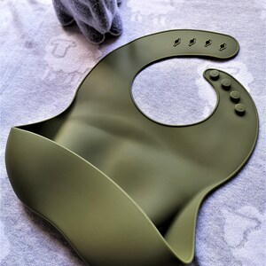 Puede incluir: Babero de silicona color caqui con un dise&ntilde;o curvo y un bolsillo para recoger la comida. El babero tiene cierres de cuello ajustables y una forma redondeada. Un peluche gris con orejas largas est&aacute; en el fondo.