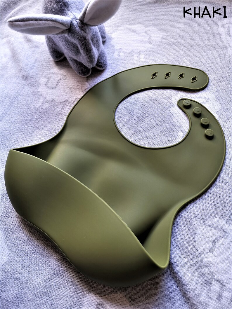 Puede incluir: Babero de silicona color caqui con un dise&ntilde;o curvo y un bolsillo para recoger la comida. El babero tiene cierres de cuello ajustables y una forma redondeada. Un peluche gris con orejas largas est&aacute; en el fondo.