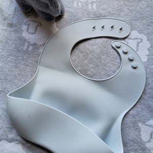 Puede incluir: Un babero de silicona azul beb&eacute; con un dise&ntilde;o curvo y un bolsillo para recoger la comida. El babero tiene broches ajustables en el cuello y se muestra sobre una superficie gris estampada. El texto "BABY BLUE" es visible.