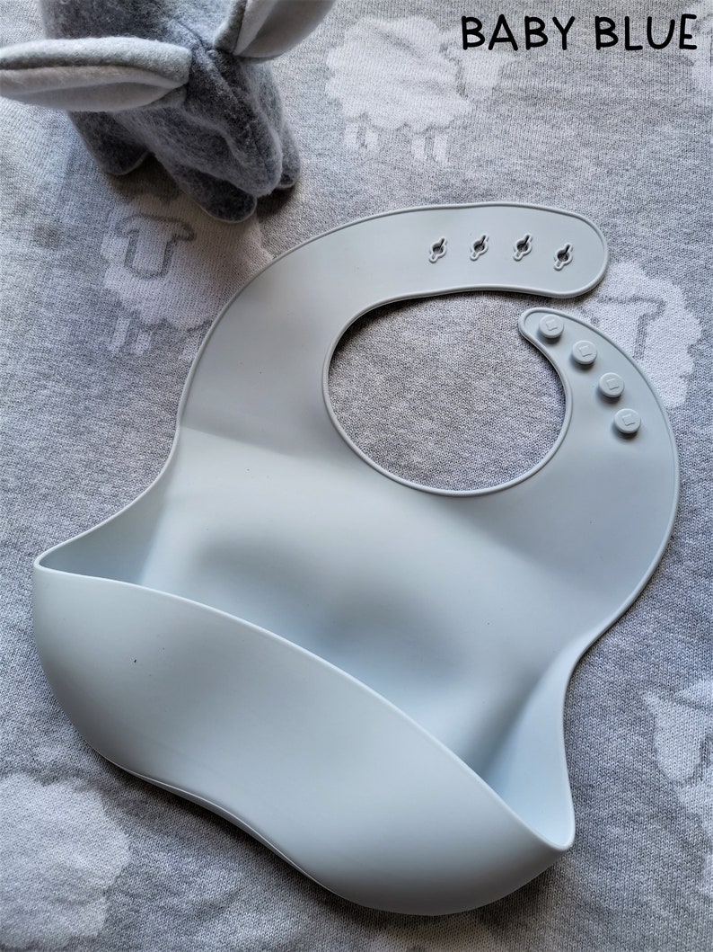 Puede incluir: Un babero de silicona azul beb&eacute; con un dise&ntilde;o curvo y un bolsillo para recoger la comida. El babero tiene broches ajustables en el cuello y se muestra sobre una superficie gris estampada. El texto "BABY BLUE" es visible.