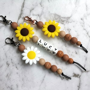 Puede incluir: Tres collares de dentición de silicona con cuentas de madera y abalorios. Los collares presentan un girasol, una margarita y el nombre "LUCY" escrito en letras blancas en cuentas individuales. Cada collar tiene un broche plateado.