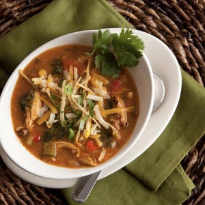 May include: A white oval bowl filled with a hearty soup, garnished with fresh cilantro, shredded cheese, and crispy tortilla strips. The soup contains visible pieces of chicken, vegetables, and corn. The bowl sits on a white plate and a green napkin, with a woven background.