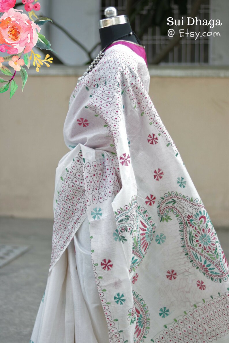Puede incluir: Sari blanco con intrincados bordados en rojo, verde y azul. El sari presenta dise&ntilde;os florales y de cachemira, con el texto "Sui Dhaga @ Etsy.com" en la esquina superior derecha. El sari est&aacute; drapeado sobre un maniqu&iacute;.