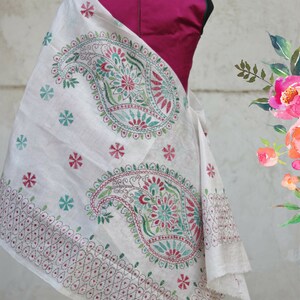 Puede incluir: Chal blanco con intrincados bordados de cachemir y flores en tonos verdes, rojos y turquesas. El chal est&aacute; drapeado sobre un maniqu&iacute; con una parte superior magenta. El nombre de la tienda Etsy "Sui Dhaga" es visible.