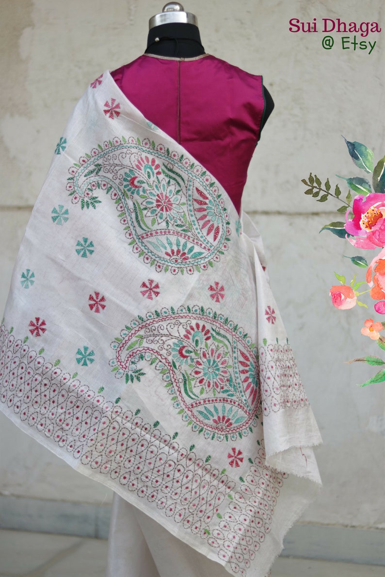 Puede incluir: Chal blanco con intrincados bordados de cachemir y flores en tonos verdes, rojos y turquesas. El chal est&aacute; drapeado sobre un maniqu&iacute; con una parte superior magenta. El nombre de la tienda Etsy "Sui Dhaga" es visible.