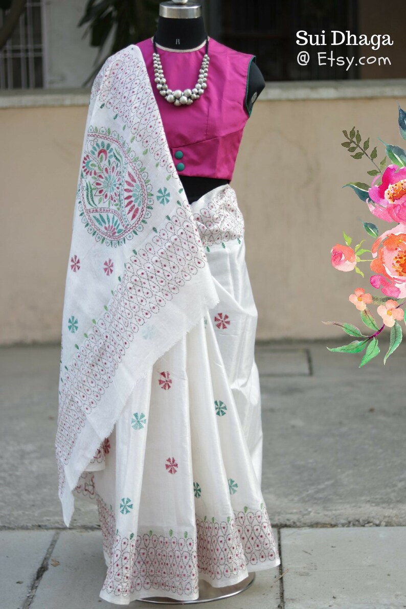 Puede incluir: Un sari blanco con intrincados bordados en patrones florales rojos, verdes y turquesas. El sari se combina con una blusa fucsia y un collar de cuentas plateadas. El texto "Sui Dhaga @ Etsy.com" es visible en la esquina superior derecha.