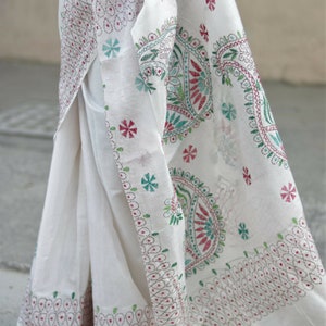 Puede incluir: Sari blanco con intrincados bordados en rojo, verde y turquesa. La tela presenta detallados patrones florales y de cachemira, con el texto "Sui Dhaga @ Etsy.com". El sari est&aacute; drapeado, mostrando el dise&ntilde;o.