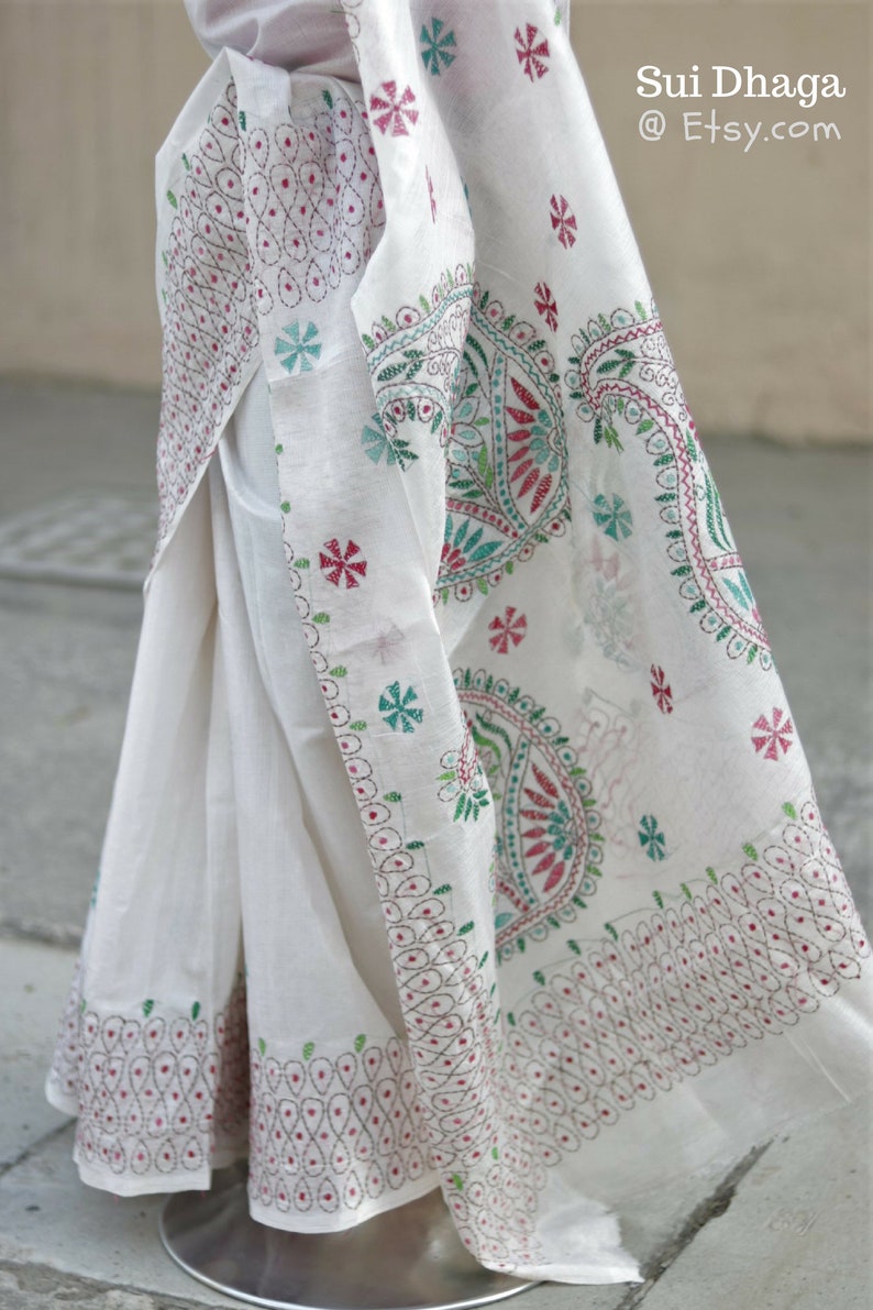Puede incluir: Sari blanco con intrincados bordados en rojo, verde y turquesa. La tela presenta detallados patrones florales y de cachemira, con el texto "Sui Dhaga @ Etsy.com". El sari est&aacute; drapeado, mostrando el dise&ntilde;o.